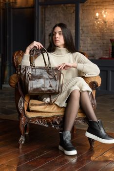 a brunette girl in a knitted beige dress poses while sitting with a shiny leather bag in her hands