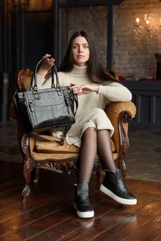 a brunette girl in a knitted beige dress poses while sitting with a shiny black leather bag in her hands