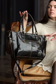 a brunette girl in a knitted beige dress poses while sitting with a shiny black leather bag in her hands