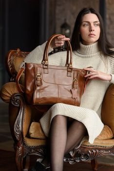 a brunette girl in a knitted beige dress poses while sitting with a shiny leather bag in her hands