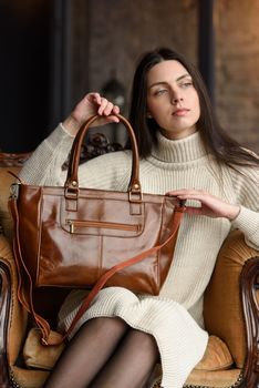 a brunette girl in a knitted beige dress poses while sitting with a shiny leather bag in her hands