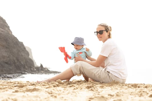 Mother playing his infant baby boy son on sandy beach enjoying summer vacationson on Lanzarote island, Spain. Family travel and vacations concept