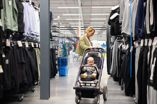 Casualy dressed mother choosing sporty shoes and clothes products in sports department of supermarket store with her infant baby boy child in stroller