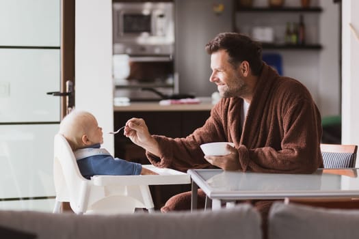 Father wearing bathrope spoon feeding hir infant baby boy child sitting in high chair at the dining table in kitchen at home in the morning