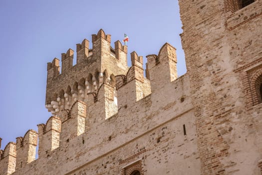 A stunning view of the castle in Sirmione, Italy, surrounded by the beautiful blue waters of Lake Garda. The ancient walls and towers of the castle create an impressive contrast against the blue sky