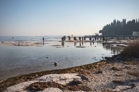 Sirmione, Italy 15 February 2023: A stunning landscape photo of a rocky beach in Sirmione, with the clear waters of Lake Garda in the background