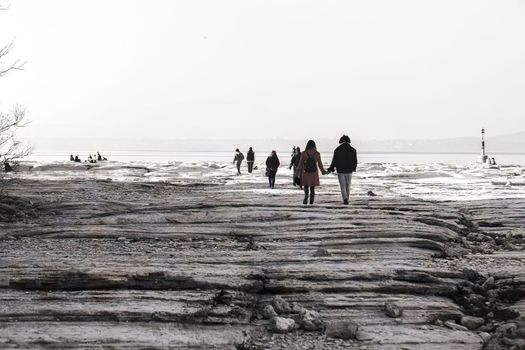 Sirmione, Italy 15 February 2023: A nostalgic shot in black and white, featuring people walking on a rocky shoreline by a lake. The dreamy atmosphere of the photo evokes a sense of longing for simpler times.