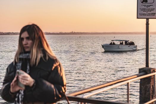 Sirmione, Italy 15 February 2023: A young woman takes a selfie on the pier with a beautiful boat in the background, against the orange and pink hues of the sunset sky.