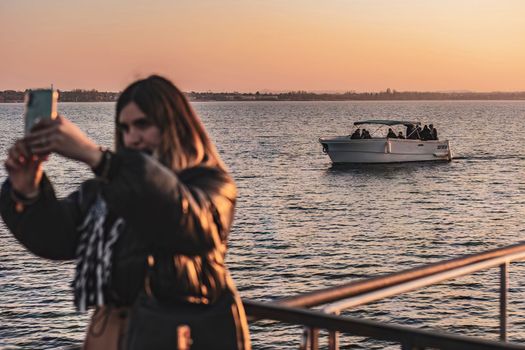 Sirmione, Italy 15 February 2023: A young woman takes a selfie on the pier with a beautiful boat in the background, against the orange and pink hues of the sunset sky.