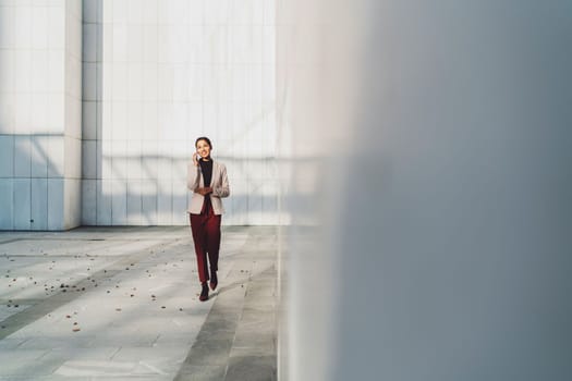 Young businesswoman walking outside while on her phone, talking to a friend on her lunch break. Business woman outside the office building on a sunny day.