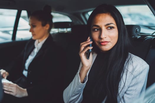 Two business women sitting in the back of a car, focus on one woman looking out the window while on a phone call.