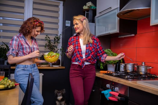 Two women styled in pin-up style stand in the kitchen cooking a home-cooked meal. On the floor sits a dog looking at a yellow bowl held by one of the women. In the background is a window with plants on the windowsill.