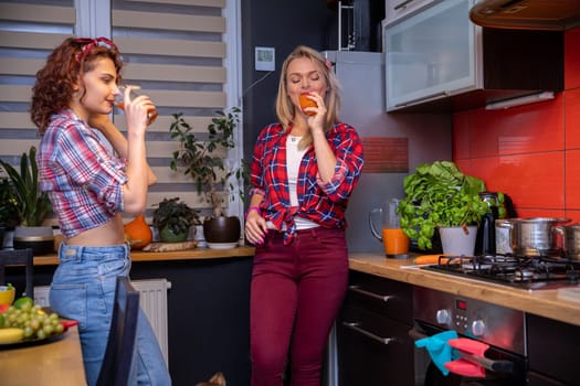 Two women dressed in pin-up style stand in the kitchen and drink fruit juice from tall glasses. There are pots on the gas stove and dishes and herbs growing in a pot on the counter next to them.