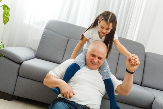 Cute little girl spending time with positive active grandpa in living room.