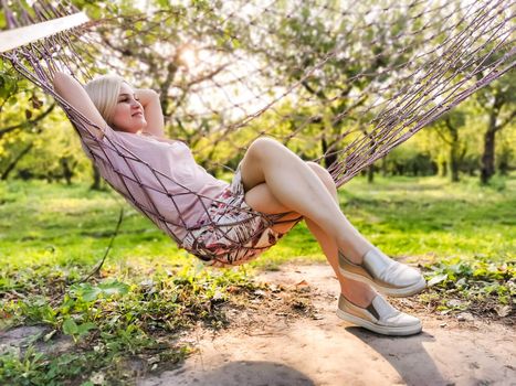 Smiling blonde woman relaxing on the hammock in garden, leisure time and summer holiday concept.