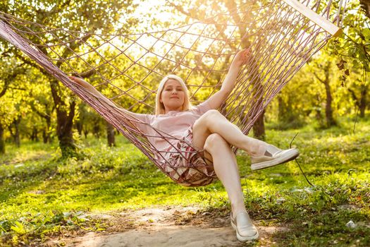 Smiling blonde woman relaxing on the hammock in garden, leisure time and summer holiday concept.