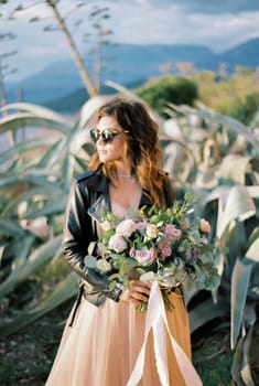 Bride in sunglasses with a bouquet of flowers stands near the agave bush. High quality photo