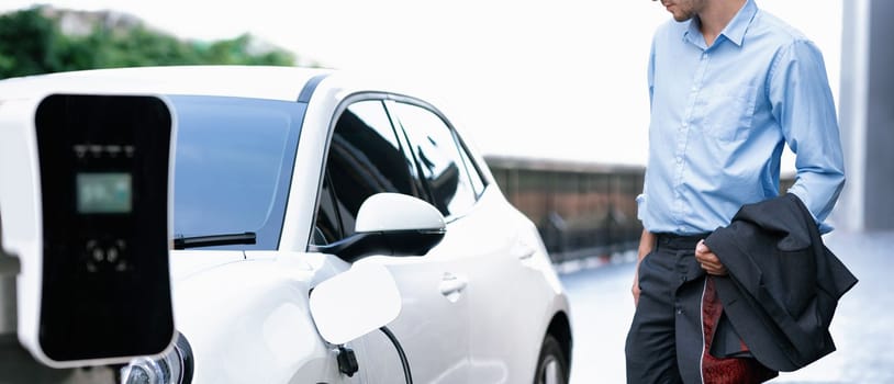 Progressive businessman with electric car recharging at public parking car charging station at modern city center. Eco friendly rechargeable car powered by alternative clean energy.