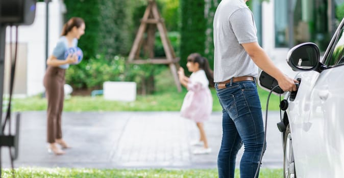 Focus image of progressive man charging electric car from home charging station with blur mother and daughter playing together in the background.
