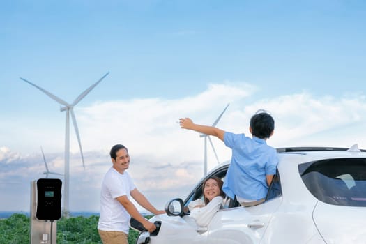 Concept of progressive happy family enjoying their time at wind farm with electric vehicle. Electric vehicle driven by clean renewable energy from wind turbine generator for charging station.