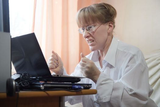 Beautiful slim blond woman in glass and white shirt with laptop computer at home near window with curtain. Emotions from online work on Internet or chat. Joy, anger, delight, surprise, indignation