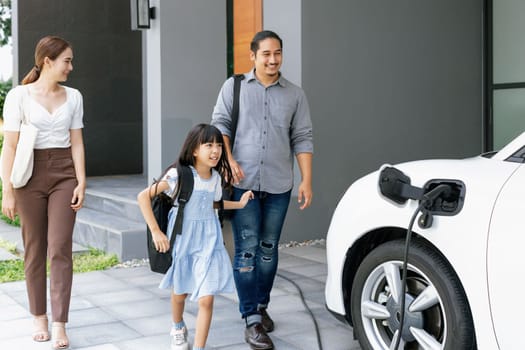 Progressive young parents and daughter with electric vehicle and home charging station. Green and clean energy from electric vehicles for healthy environment. Eco power from renewable source at home.
