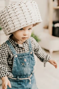Little girl 1 years old with a basket on her head. Funny child plays puts a basket on her head like a hat and laughs, she is dressed in a plaid shirt and denim overalls. Funny moments with kids