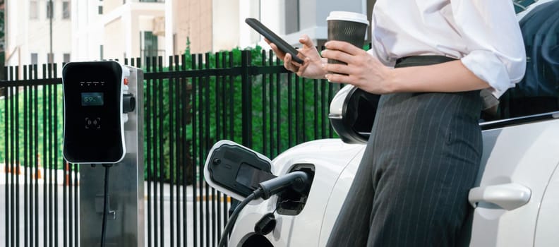 Closeup progressive suit-clad businesswoman with her electric vehicle recharge her car on public charging station in modern city with power cable plug and renewable energy-powered electric vehicle.