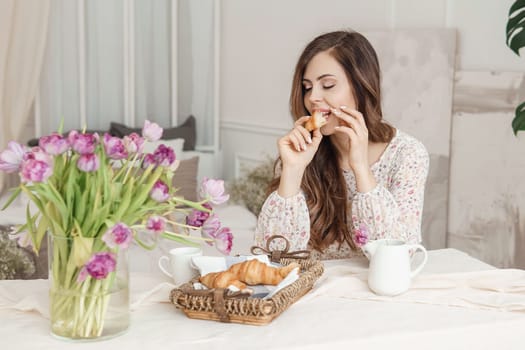 A brown-haired woman with long hair is having breakfast at the table with croissants and a cup of coffee. Spring portrait