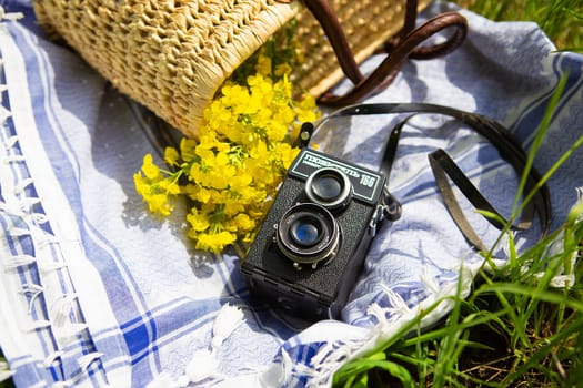 A straw picnic basket lies on a blue blanket on green grass along with a bouquet of yellow flowers. In the background is an old camera with the name Lover 166 written on it