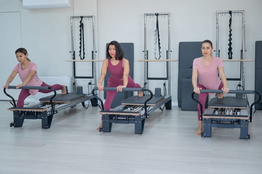 Three Asian women in pink sportswear do exercises on the reformer machine. Pilates classes