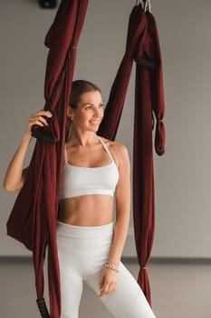 A portrait of a girl in white sportswear stands near a hanging Yoga hammock in the fitness room.