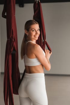 A portrait of a girl in white sportswear stands near a hanging Yoga hammock in the fitness room.
