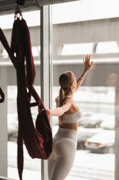 Portrait of a girl in white sportswear Standing near the window and holding a hanging hammock in her hand in the fitness room.