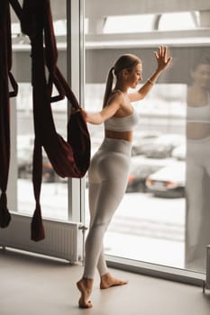 Portrait of a girl in white sportswear Standing near the window and holding a hanging hammock in her hand in the fitness room.