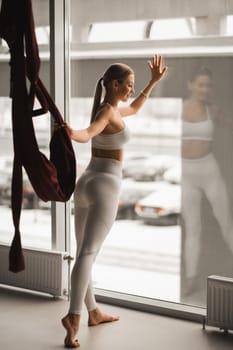 Portrait of a girl in white sportswear Standing near the window and holding a hanging hammock in her hand in the fitness room.