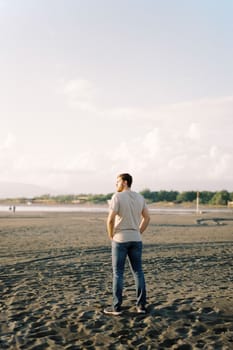 Young man in jeans stands on a sandy beach and looks at the sea. High quality photo