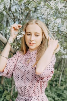 Blonde girl on a spring walk in the garden with cherry blossoms. Female portrait, close-up. A girl in a pink polka dot dress