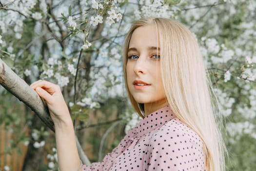 Blonde girl on a spring walk in the garden with cherry blossoms. Female portrait, close-up. A girl in a pink polka dot dress