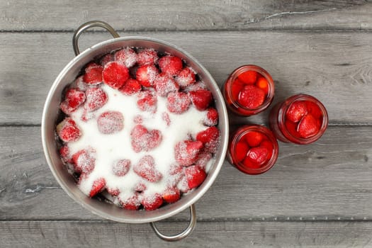 Tabletop view - steel put with strawberries covered in crystal sugar, three bottles with pickled strawberry next to it. Home made jam / compote preparation