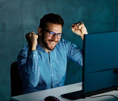 Portrait of a happy young businessman by monitor and desktop computer working late celebrating success and good news at night in office