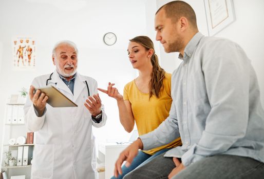 Senior doctor holding tablet talking to young couple in his office in clinic