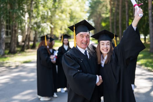 A group of graduates in robes outdoors. An elderly man and a young woman congratulate each other on their graduation