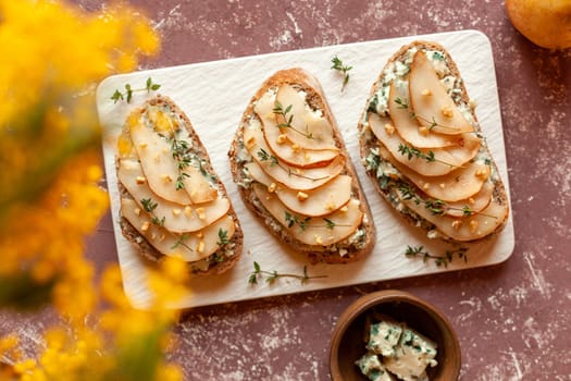 roquefort french cheese and sliced peaches toasts on a square plate, on a table under mimosa flowers, top view