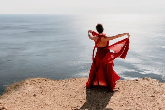 Side view a Young beautiful sensual woman in a red long dress posing on a rock high above the sea during sunrise. Girl on the nature on blue sky background. Fashion photo.