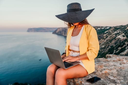 Successful business woman in yellow hat working on laptop by the sea. Pretty lady typing on computer at summer day outdoors. Freelance, travel and holidays concept.