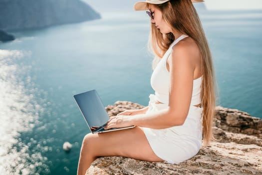 Successful business woman in yellow hat working on laptop by the sea. Pretty lady typing on computer at summer day outdoors. Freelance, travel and holidays concept.