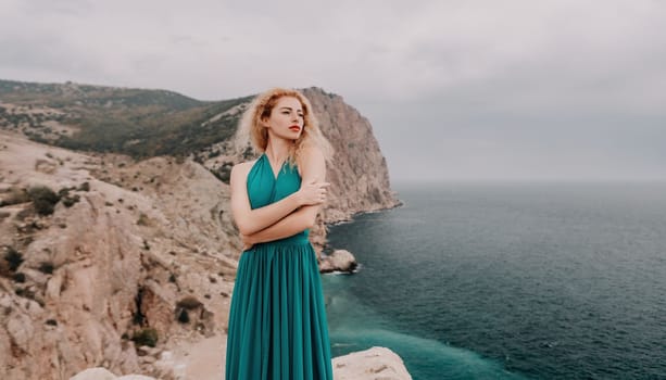 Side view a Young beautiful sensual woman in a mint long dress posing on a volcanic rock high above the sea during sunset. Girl on the nature on overcast sky background. Fashion photo