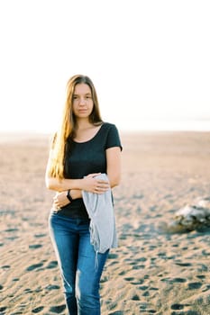 Girl in jeans with a sweater in her hands stands on a sand. High quality photo