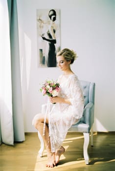Bride in a lace dressing gown with a bouquet sits in a chair. High quality photo